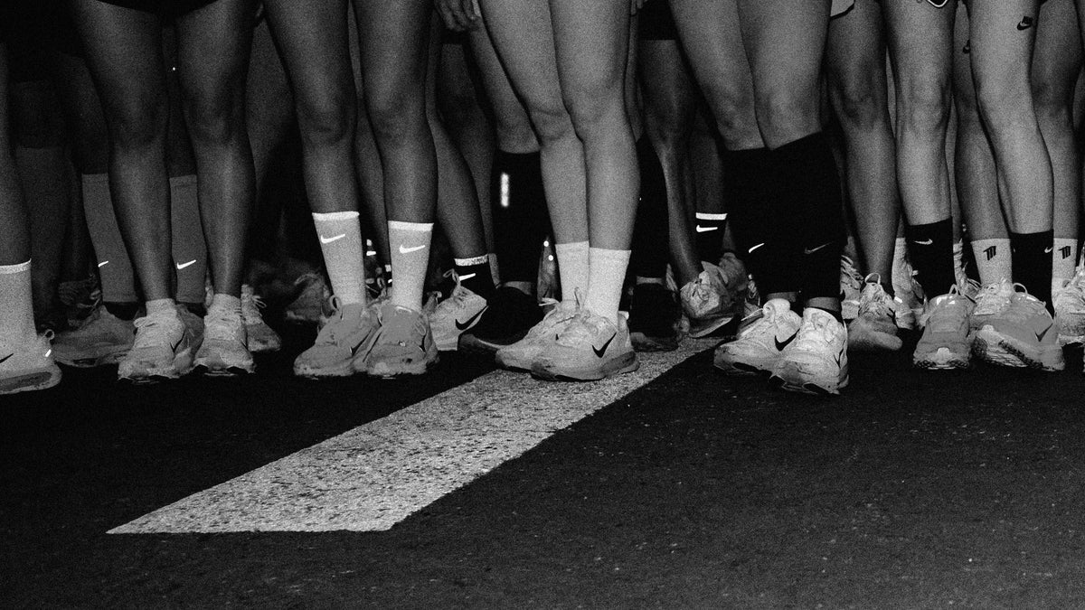 Black and white photo of multiple people's legs and feet standing together on a dark pavement surface, with white athletic socks and sneakers visible and a painted white line running beneath them.